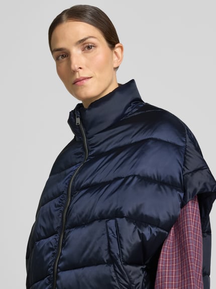 Frauenmodel trägt Steppweste mit Stehkragen und Eingrifftaschen von Milano Italy, glänzendes Dunkelblau, glatte Textur, Nahaufnahme Oberkörper, Relaxed Fit, Reißverschluss, Rückenlänge 59 cm, Schulterbreite 50 cm, Brustweite 63 cm.