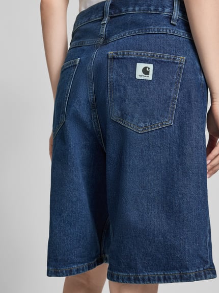 Frauenmodel trägt Relaxed Fit Jeans Shorts aus reiner bawełna Modell 'Brandon' von Carhartt Work In Progress, 5 kieszeni, naszywka z logo, zapięcie na guzik i zamek błyskawiczny, szerokość w rozmiarze S: 41 cm, długość wewnętrzna nogawki w rozmiarze S: 24 cm, widoczne ciemnoniebieskie odcienie i gładka struktura tkaniny, Ganzkörperansicht Rückansicht.