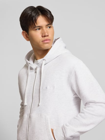 Männermodel trägt Relaxed Fit Sweatjacke aus Baumwoll-Mix Modell 'ORSALAMANCO' von JACK & JONES in Hellgrau mit Kapuze, Nahaufnahme Oberkörper. Sweater zeigt Zweiwege-Reißverschluss, Känguru-Tasche und elastische Abschlüsse, weiche Stoffstruktur.