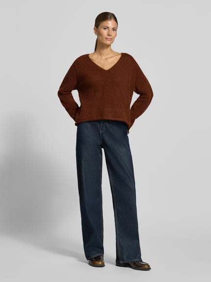 Frauenmodel trägt Relaxed Fit Strickpullover aus Alpaka-Mix Modell 'SOMAIJA' von DRYKORN in Braun, kombiniert mit Jeans, Ganzkörperansicht von vorne. Damen Strickpullover mit V-Ausschnitt, Rollsaum, Rückenlänge 55 cm, Ärmellänge 60 cm.