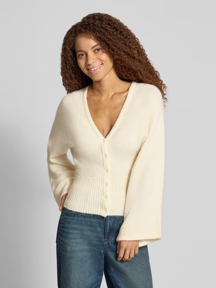 Vrouwenmodel draagt Regular Fit gebreid jack met wol van Gina Tricot, elastische materiaalmix, ribboorden, knoopsluiting, met lange mouwen, ruglengte maat S 53 cm, mouwlengte 49 cm, effen licht crème kleur, zachte textuur, voorkant bovenlichaam close-up.