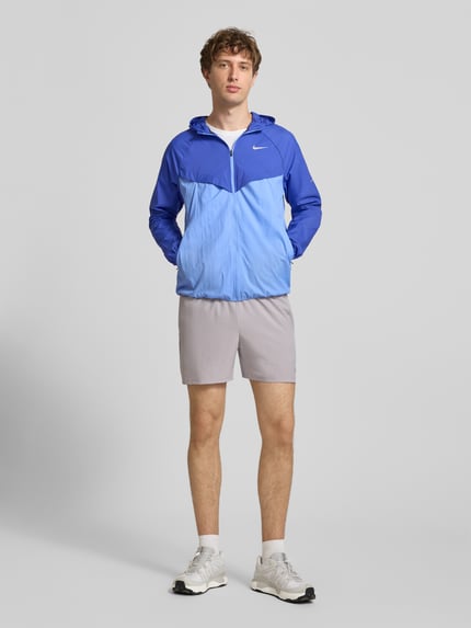 Männermodel trägt Trainingsjacke mit Logo-Print und Kapuze von NIKE TRAINING in zwei Blautönen, Reißverschluss, gerader Schnitt, Ganzkörperansicht von vorne.