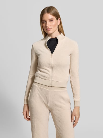 Frauenmodel trägt Slim Fit Sweatjacke mit Stehkragen Modell 'LAUDOMIA' von GUESS, beigefarbener Material-Mix mit Logo-Muster, Ganzkörperansicht von vorne.