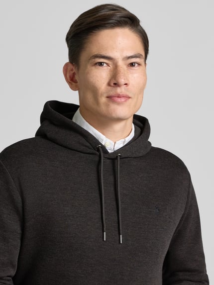 Męski model nosi Hoodie z kapturem marki Polo Ralph Lauren w kolorze ciemnoszarym, bawełniany miks, luźny krój, zbliżenie na górną część ciała.