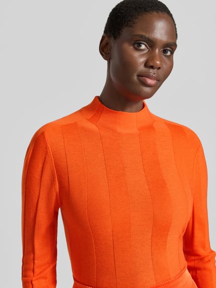 Frauenmodel trägt Strickpullover aus Schurwolle-Mix mit Stehkragen von Marc Cain in Orange, Slim Fit, feine Struktur, Nahaufnahme Oberkörper. Damenpullover mit Kaschmir-Anteil, Rückenlänge 55 cm, Ärmellänge 66 cm, Label-Detail sichtbar.