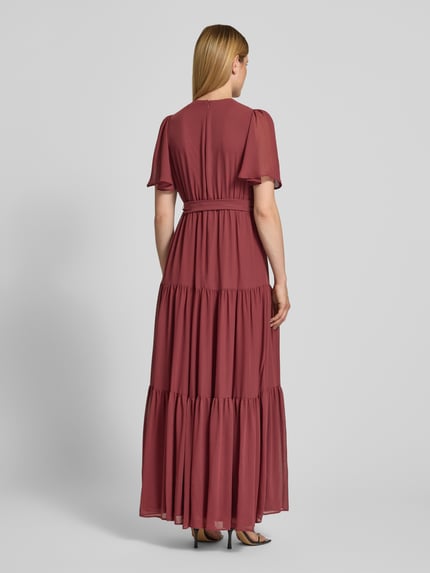 Frauenmodel trägt Abendkleid mit V-Ausschnitt und Bindegürtel von Christian Berg Cocktail aus Chiffon, Stufen-Look, A-Linie, Maxikleid, Rückenlänge bei Größe 36: 151 cm, Brustweite 41 cm, Ärmellänge 30 cm, Ganzkörperansicht Rückansicht.