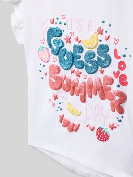 Meisje draagt Regular Fit T-Shirt met Statement-Print van Guess, wit katoenmix met kleurrijke motief- en logo-opdruk, ruches, detailopname.