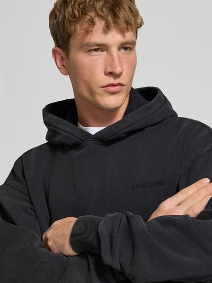 Männermodel trägt Oversized Hoodie aus Baumwoll-Mix von PEGADOR in Schwarz mit Logo-Stickerei, Nahaufnahme Oberkörper.