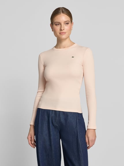 Frauenmodel trägt Slim Fit Langarmshirt aus reiner Baumwolle von Lacoste in zartem Rosa, kombiniert mit dunkler Hose, Nahaufnahme Oberkörper, Damen T-Shirt von Lacoste.