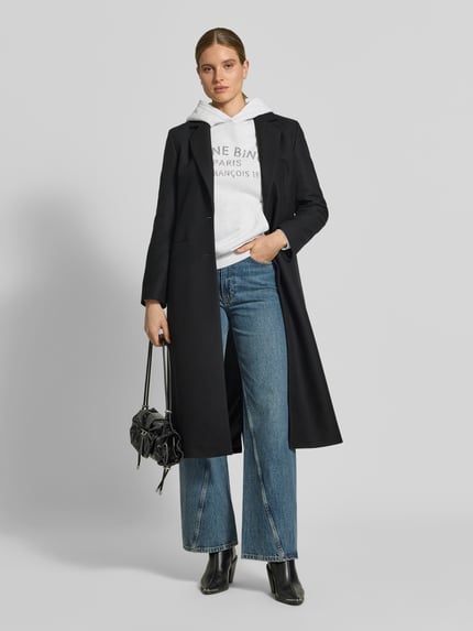 Frauenmodel trägt Oversized Hoodie aus reiner Baumwolle von Anine Bing in Grau mit Label-Print und Känguru-Tasche zu Jeans, Ganzkörperansicht von vorne.