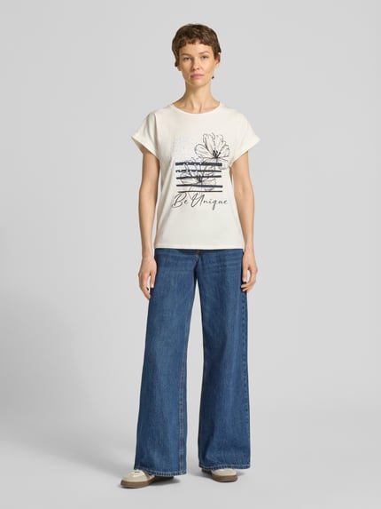 Vrouwenmodel draagt T-shirt met fixeerde mouwomslag model 'RINA' van Soyaconcept, recht model met ronde hals, motiefprint, katoenmix met stretch, crèmekleurig, gecombineerd met blauwe jeans, frontale opname van het hele lichaam.