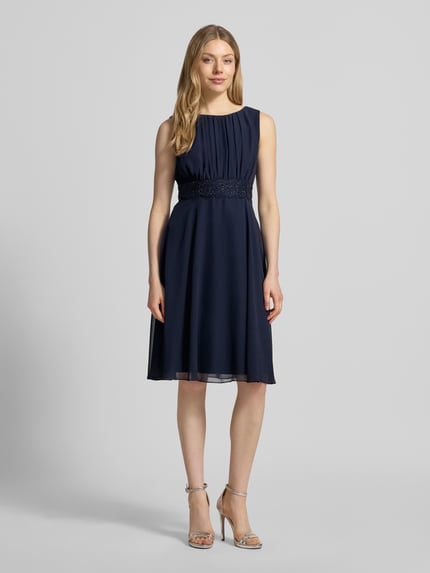 Frauenmodel trägt Knielanges Cocktailkleid mit Taillenband von Christian Berg Cocktail, dunkelblauer Chiffon, Drapierungen, taillierter Schnitt, Rundhalsausschnitt, Knielänge, Ganzkörperansicht von vorne. Rückenlänge bei Größe 36: 103 cm, Brustweite bei Größe 36: 36 cm.