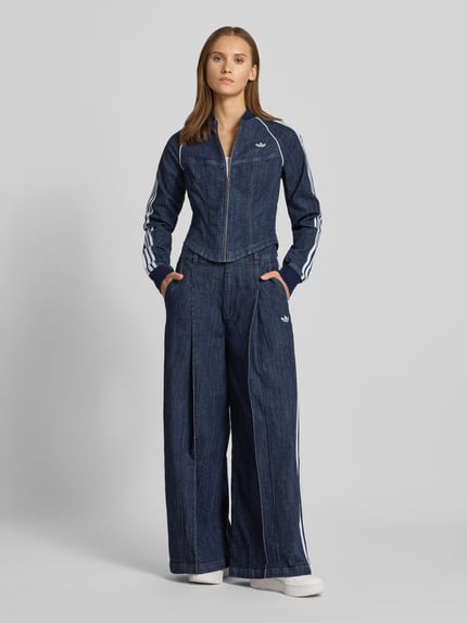 Modelka kobieca nosi Regular Fit Jeansjacke aus Baumwoll-Mix Modell 'SUPERSTAR' od adidas Originals, ciemnoniebieska kurtka jeansowa z białymi paskami i teksturą denimu, pełny widok z przodu.