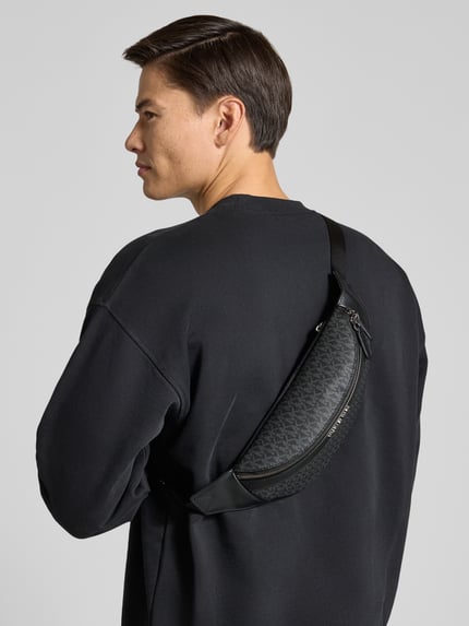 Männermodel trägt Bauchtasche mit Logo-Muster Modell 'GREYSON' von Michael Kors in Schwarz, Detailansicht Rückansicht. Herren Bauchtasche mit Hauptfach und Reißverschluss, Allover-Look, Schulterriemen ca. 82 cm, Maße ca. 28x15x10 cm.