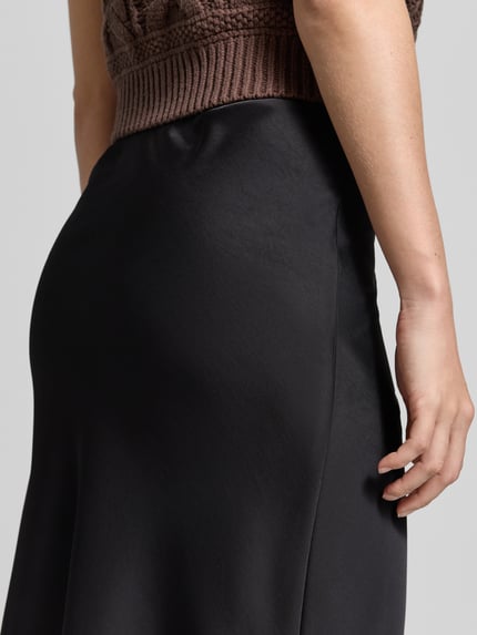 Frauenmodel trägt Satinrock mit elastischem Bund Modell 'Mindie' von Neo Noir, glatter schwarz glänzender Stoff, Detailaufnahme, Material-Mix mit Stretch-Anteil, Straight Fit, mittellanger Schnitt.