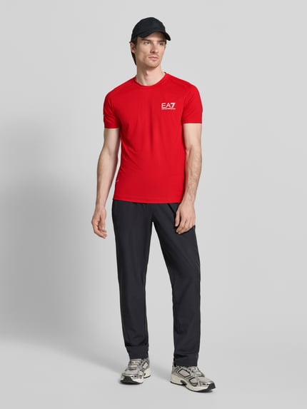 Męski model nosi T-Shirt mit Label-Print od EA7 Emporio Armani, czerwony kolor, krój Slim Fit, widok całej sylwetki z przodu.