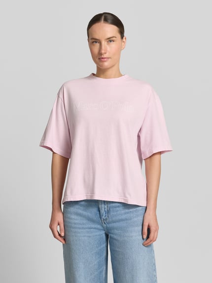 Kobiece modelka nosi Boxy Fit T-Shirt aus reiner Baumwolle von Marc O'Polo, jednokolorowy, pudełkowy krój, aplikacja: z napisem z logo, okrągły dekolt, krótki rękaw, materiał: bawełna, jasnoróżowy, gładka tekstura, pełen widok z przodu.