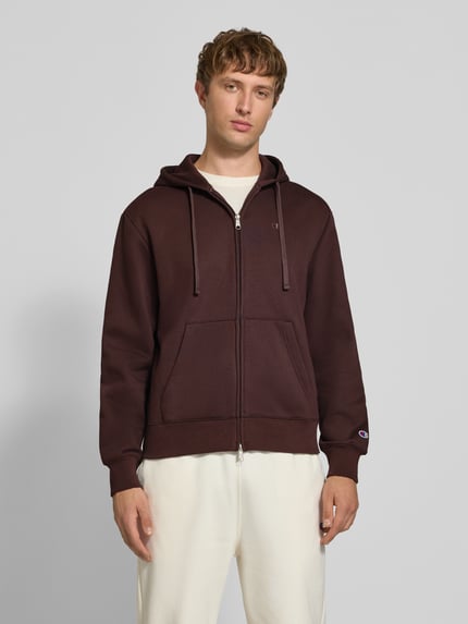 Männermodel trägt Sweatjacke mit Label-Stitching und Känguru-Tasche von Champion im Baumwoll-Mix in Dunkelbraun, Ganzkörperansicht von vorne.