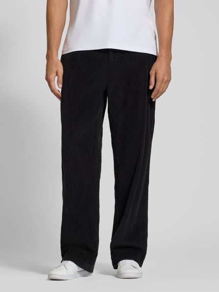 Männermodel trägt schwarze Cordhose mit Struktur, weiter Schnitt, langer Beinverlauf, Kontrast durch weiße Sneaker, Nahaufnahme Unterkörper.