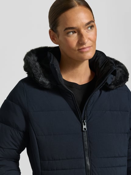 Frauenmodel trägt Jacke mit abnehmbarer Kapuze Modell 'ECLIPSE' von NAUTGÅRD in Dunkelblau, gesteppt, mit Reißverschluss, Nahaufnahme Oberkörper. Rückenlänge 79 cm, Schulterbreite 38 cm, Brustweite 48 cm, Ärmellänge 62 cm, gerader Schnitt, seitliche Taschen, gestrickte Ärmelabschlüsse.