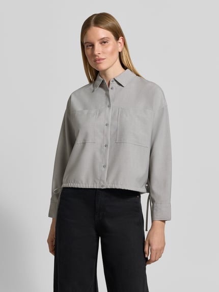 Frauenmodel trägt Cropped Langarmbluse mit Tunnelzugsaum Modell 'Felanga' von OPUS, graue Melange-Optik mit Struktur, Knöpfe, Kordeln, Brusttasche, Webware, Ganzkörperansicht von vorne.
