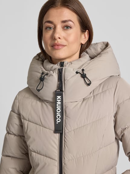 Kobiece model nosi kurtkę pikowaną z kapturem i detalem z logo Model 'HERSCH2' od khujo, beżowy kolor, pikowanie, zamek błyskawiczny, z dzianinowymi zakończeniami rękawów, z kieszeniami na zamek, prosty krój, z tunelowym ściągaczem w kapturze, szerokość w rozmiarze S: 50 cm, długość tyłu: 63 cm, szerokość ramion: 44 cm, długość rękawów: 64 cm, zbliżenie na górną część ciała