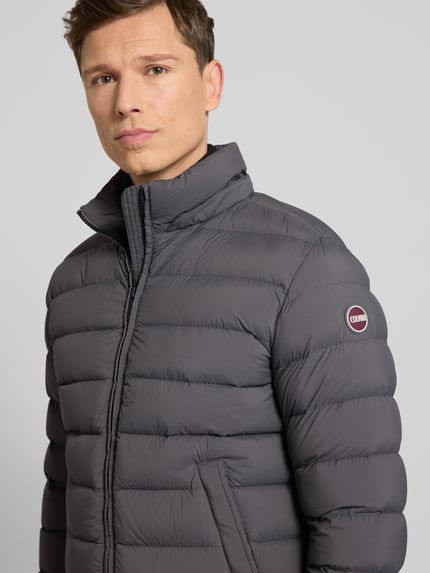 Męski model nosi Kurtkę pikowaną z kapturem od Colmar Originals, krój regular fit, zamek błyskawiczny, wpuszczane kieszenie, kolor szary, długość tyłu 66 cm, szerokość ramion 42 cm, szerokość klatki piersiowej 56 cm, długość rękawów 66 cm, detale poziomych pikowań; nahaufnahme górnej części ciała.