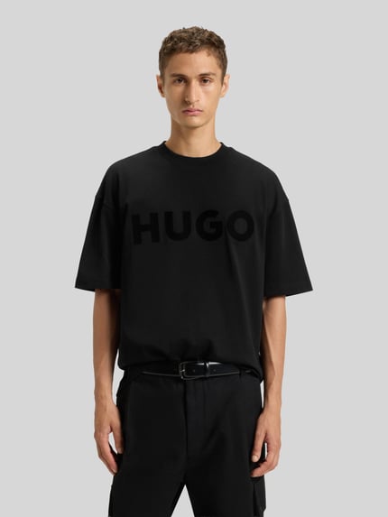 Męski model nosi Oversized T-Shirt aus reiner Baumwolle Modell 'DINKEE' od HUGO, czarne, gładkie, nadruk z logo, okrągły dekolt, krótki rękaw, bawełna, Ganzkörperansicht von vorne.