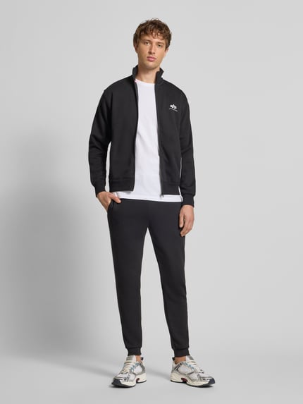 Männermodel trägt Sweatjacke mit Logo und Stehkragen von Alpha Industries aus Baumwoll-Mix, Regular-Fit, schwarz, Ganzkörperansicht von vorne.