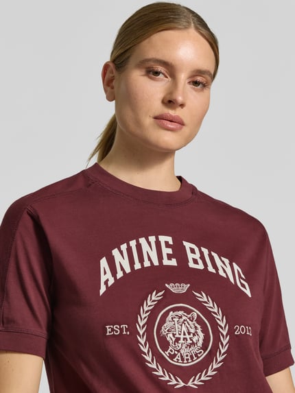 Modelka kobieca nosi bordowe T-Shirt mit Label-Print od Anine Bing z białym nadrukiem, czysta bawełna, krój regular fit, okrągły dekolt, długość tyłu w rozmiarze S: 58 cm, zbliżenie na górną część ciała.