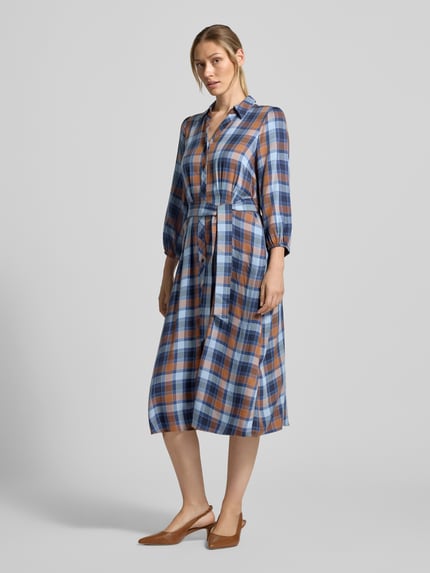 Frauenmodel trägt Midikleid aus reiner Viskose mit Umlegekragen von MORE & MORE, kariertes Muster in Blau-Braun, Ganzkörperansicht von vorne. Taillierte Passform, durchgängige Knopfleiste, 3/4-Arm mit elastischen Abschlüssen, Taillenband, Rückenlänge 112 cm, Brustweite 34 cm, Ärmellänge 51 cm.