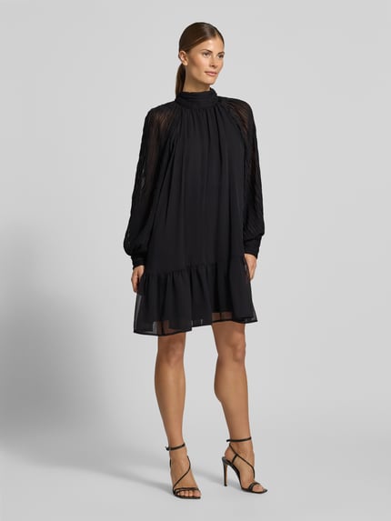 Frauenmodel trägt Minikleid mit Stehkragen Modell 'OLERA' von YAS, A-Linie, fließender schwarzer Stoff, Ganzkörperansicht von vorne.