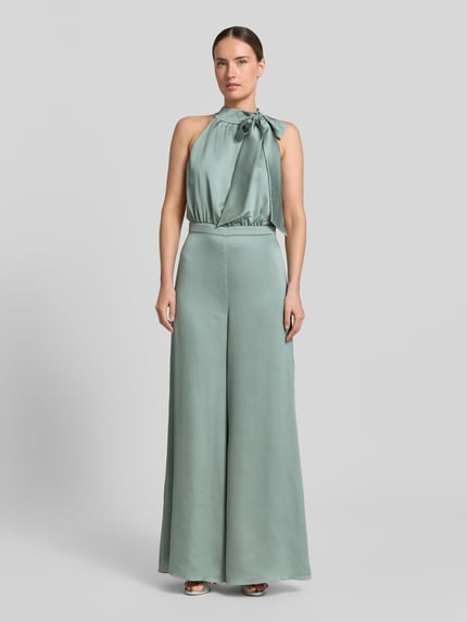 Modelka kobieca ma na sobie Jumpsuit mit Schluppe und rückseitigem Knopfverschluss od Swing, satyna, krój slim fit, kolor pastelowozielony, całkowita długość 152 cm, długość wewnętrzna nogawki 82 cm, szerokość w rozmiarze 36: 66 cm, z tyłu zapięcie na guzik oraz zatrzask i zamek, widok całej sylwetki z przodu.