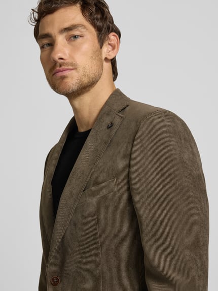Männermodel trägt Sakko mit Pattentaschen und Reverskragen Modell 'Theo' von Carl Gross aus braunem Cord, Nahaufnahme Oberkörper. Regular Fit, sichtbare Knopfleiste und Brustleistentasche mit Einstecktuch, strukturierte Oberfläche.