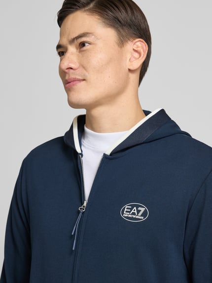 Männermodel trägt Sweatjacke mit Logo und Zweiwege-Reißverschluss von EA7 Emporio Armani in Dunkelblau, Viskose-Mix mit Stretch-Anteil, Regular Fit, Kapuze, Eingrifftaschen, Nahaufnahme Oberkörper.