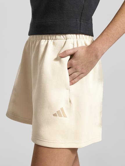 Frauenmodel trägt Shorts mit elastischem Bund von adidas Sportswear in Beige, lockerer Schnitt, Label-Print, Nahaufnahme Oberschenkel.