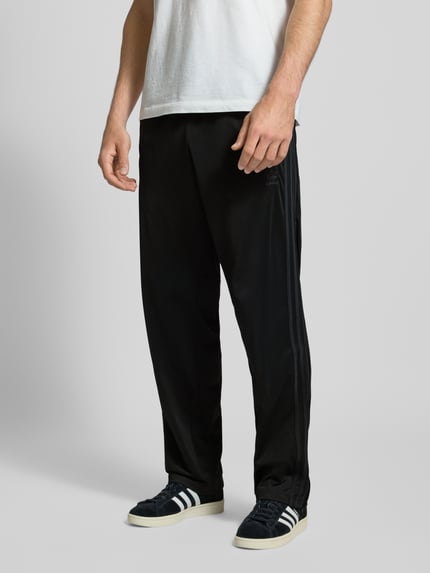 Männermodel trägt czarne spodnie treningowe Trainingshose mit elastischem Bund Modell 'FIREBIRD' von adidas Originals, Regular Fit, z kieszeniami na zamek, długość wewnętrzna nogawki 43 cm, widok na całe nogi z przodu.