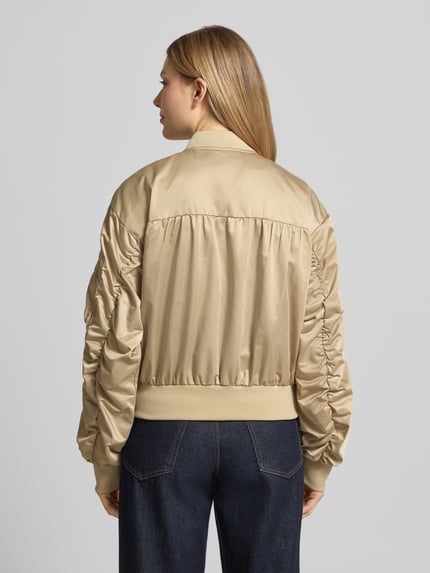 Frauenmodel trägt Bomberjacke mit Reißverschlusstasche am Ärmel von Original Bombers in Beige, Lockerer-Schnitt, Rückenansicht. Rückenlänge 49 cm, Schulterbreite 50 cm, Brustweite 58 cm, Ärmellänge 61 cm, Stehkragen, Reißverschluss, Logo-Stitching, Seitliche Eingrifftaschen mit Druckknopfverschluss, Label-Patch, Elastische Abschlüsse.