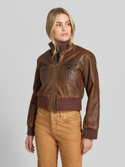 Frauenmodel trägt Lederjacke Modell 'BAILEY' mit Stehkragen von BAUM & PFERDGARTEN in brauner Lederoptik, Used Look, strukturierte Oberfläche, Ziernähte, metallische Akzente, Zierknöpfe, Label-Detail, Brusttasche, Eingrifftaschen, Zweiwege-Reißverschluss, gerippte Abschlüsse, Regular Fit, cropped, Ganzkörperansicht von vorne.