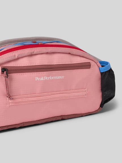Produktfoto, Bauchtasche mit Label-Detail von PEAK PERFORMANCE in Rosa, Rot und Blau, Hauptfach mit Reißverschluss, Nebenfächer, verstellbarer Riemen, Innenfächer, Breite 41 cm, Länge 18 cm, Tiefe 7 cm, Riemenlänge 65 cm, Stoff mit glatter Textur.