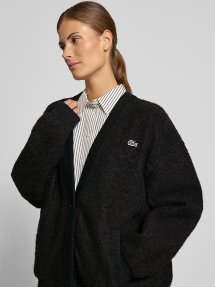 Frauenmodel trägt Cardigan mit Logo-Patch von Lacoste, melierte Strickoptik, V-Ausschnitt, Ripprand, Knöpfe, Nahaufnahme Oberkörper.
