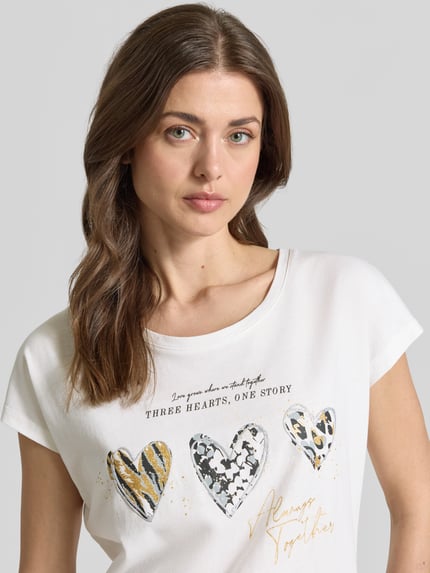 Modelka nosi Regular Fit T-Shirt mit Viskose-Anteil od Christian Berg Woman, biały, nadruk z motywem, bawełniany miks, krótki rękaw, okrągły dekolt, przód z napisem i sercami, krój regular fit, widok na górną część ciała z przodu.