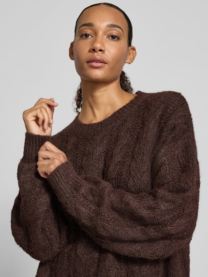 Frauenmodel trägt Oversized Fit Strickpullover mit Zopfmuster von REVIEW in Dunkelbraun, sichtbarer Rundhalsausschnitt, Nahaufnahme Oberkörper. Rückenlänge 54 cm, Ärmellänge 58 cm, elastische Abschlüsse.