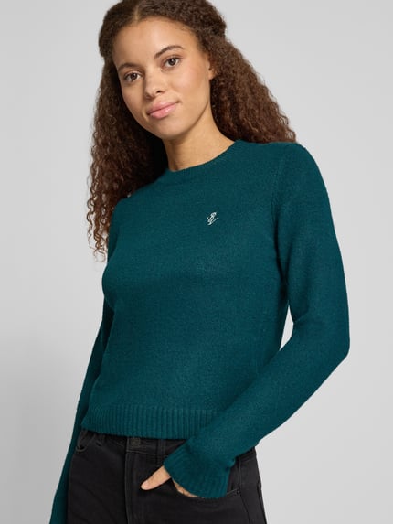 Modelka nosi Slim Fit Strickpullover aus elastischem Material-Mix von REVIEW, sweter z dzianiny, elastyczna mieszanka materiałów, krój slim fit, Wyhaftowany motyw, prążkowane wykończenia, długość tyłu w rozmiarze S: 51 cm, długość rękawów w rozmiarze S: 61 cm, kolor morski, prosty fason, jednolita tekstura, widoczne haftowane logo z przodu, widoczne prążkowania na wykończeniach, Nahaufnahme górnej części ciała.
