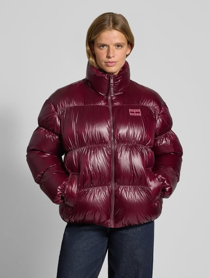 Frauenmodel trägt Regular Fit Steppjacke mit Label Stitching von Tommy Jeans in glänzendem Dunkelrot, Ganzkörperansicht von vorne.
