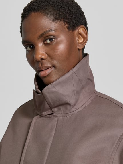 Frauenmodel trägt Jacke aus reiner Baumwolle von Victoria Beckham in Braun, glatte Textur, Stehkragen, Nahaufnahme Oberkörper. Regular Fit, Eingrifftaschen, elastische Ärmelabschlüsse, Rückenlänge bei Größe 36: 54 cm, Brustweite 50 cm, Ärmellänge 63 cm.
