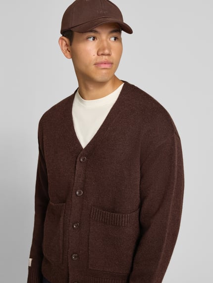 Männermodel trägt Cardigan mit rückseitigem Motiv und V-Ausschnitt von ANOTHER COTTON LAB in Braun, gerade Form, aufgesetzte Taschen, Nahaufnahme Oberkörper.