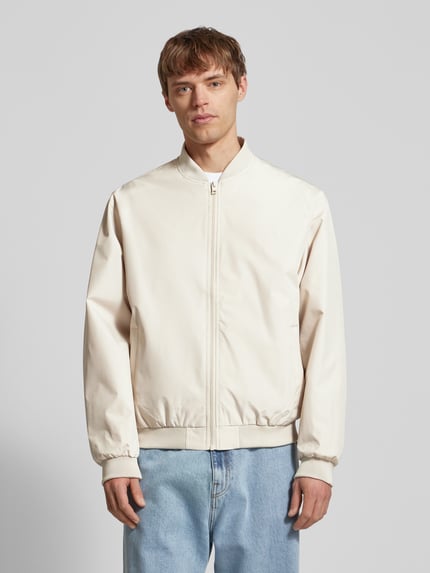 Męski model nosi bomberkę z bocznymi kieszeniami model 'CHARGE' od Jack & Jones, krój straight fit, kremowy, zamek, stójka, prążkowane wykończenia, długość tyłu w rozmiarze M: 66 cm, szerokość ramion w rozmiarze M: 54 cm, szerokość klatki piersiowej w rozmiarze M: 65 cm, długość rękawa w rozmiarze M: 64 cm, widok całej sylwetki z przodu.