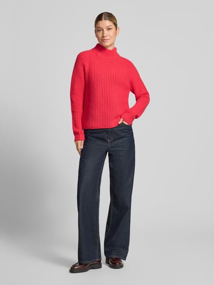 Frauenmodel trägt Strickpullover aus Kaschmir-Baumwoll-Mix Modell 'LONA' von 0039 Italy in Rot, kombiniert mit dunkler Jeans, klar strukturierte Stricktextur, Ganzkörperansicht von vorne, Regular Fit, Stehkragen, Rückenlänge 60 cm, Ärmellänge 78 cm.
