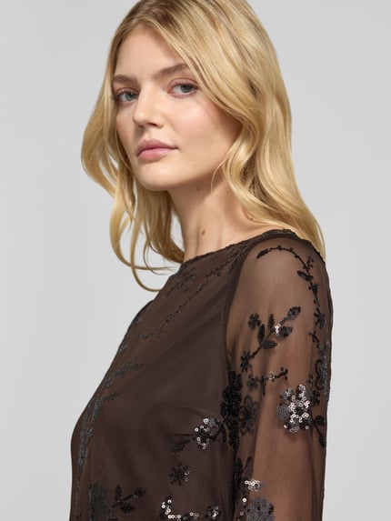 Frauenmodel trägt Kurz Langarmbluse mit Paillettenapplikation von PIECES, transparentes Chiffon mit floralem Spitzenmuster, Nahaufnahme Oberkörper.